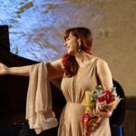 Anna Maria Chiuri, mezzosoprano, durante un concerto dal vivo, in abito da sera color oro con bouquet di fiori in mano, mentre ringrazia il pubblico accanto a un pianoforte.